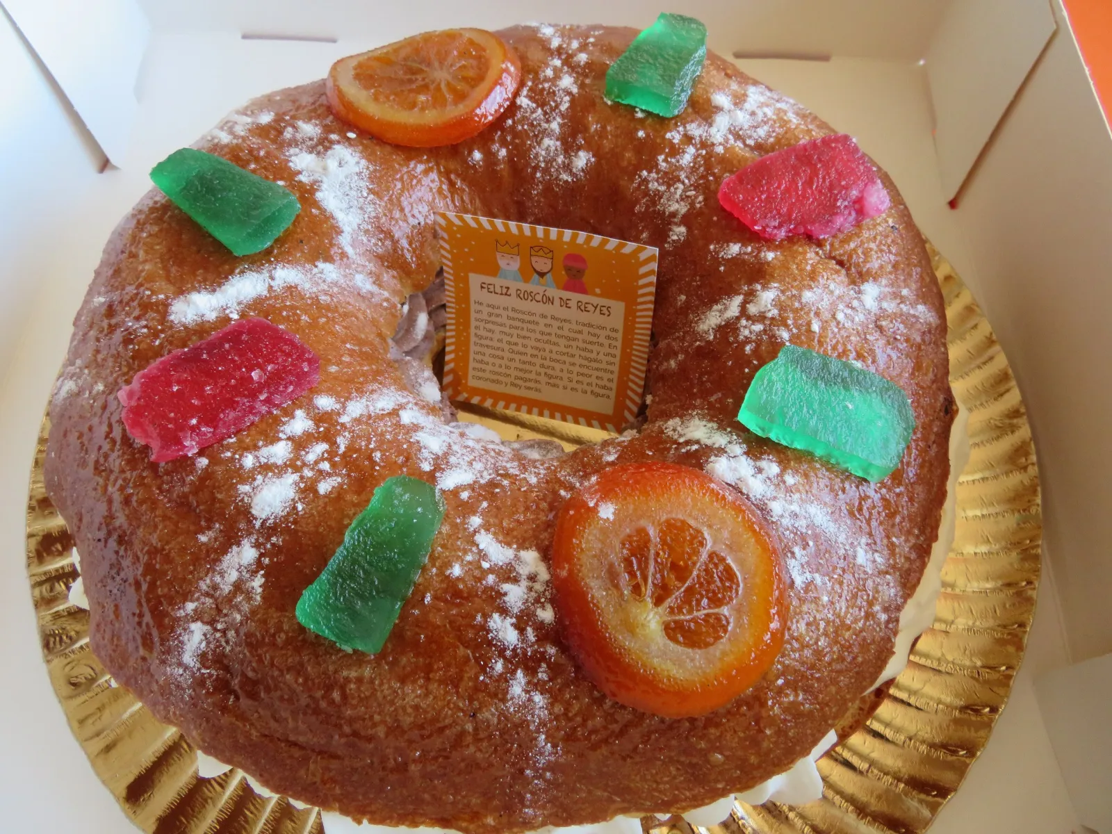 Roscón de Reyes decorado con rodajas de naranja confitada y fruta escarchada, espolvoreado con azúcar, en su caja de pastelería